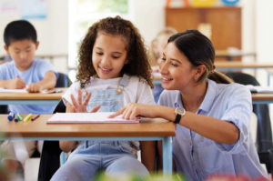 Teacher works with young girl at her desk in classroom